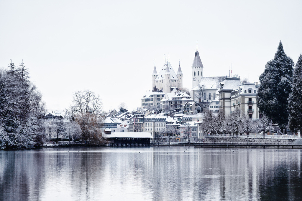 Winterliche Stadt Thun | Die historische Stadt Thun präsentiert sich in einer winterlichen Schneelandschaft, mit dem imposanten Schloss Thun und der Stadtkirche als zentrale Elemente. Die verschneiten Gebäude und Bäume spiegeln sich ruhig im Wasser des Sees wider, was eine friedliche und malerische Szene schafft. - Realisiert mit Pictrs.com