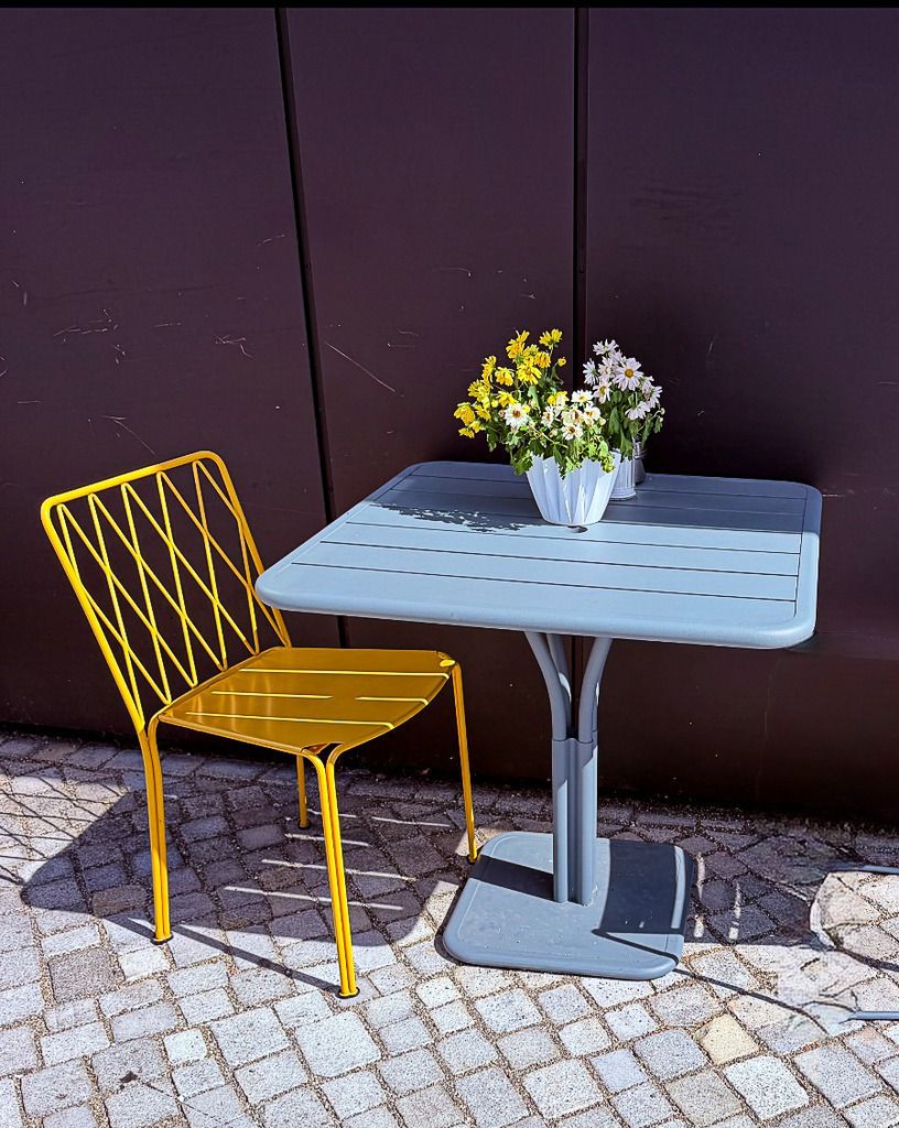 Urbane Café-Momente – Gelber Designstuhl und Blumen-Idylle | Dieses stilvolle Fotokunst-Wandbild fängt die friedliche Atmosphäre eines urbanen Cafés ein. Der leuchtend gelbe Metallstuhl und der mattblaue Tisch bilden ein harmonisches Farbduo, abgerundet durch ein frisches Blumengesteck. Die detailreichen Texturen des Pflasterbodens und der dunklen Wand verleihen dem Motiv eine spürbare Haptik. Fotografiert von Robert Steinke. Unsere Empfehlung: Als hochwertiges Alu-Dibond oder hinter glänzendem Acrylglas kommen die Kontraste und Oberflächenstrukturen in Galerie-Qualität atemberaubend zur Geltung. Auch als klassische Leinwand oder Poster verfügbar. - Realisiert mit Pictrs.com