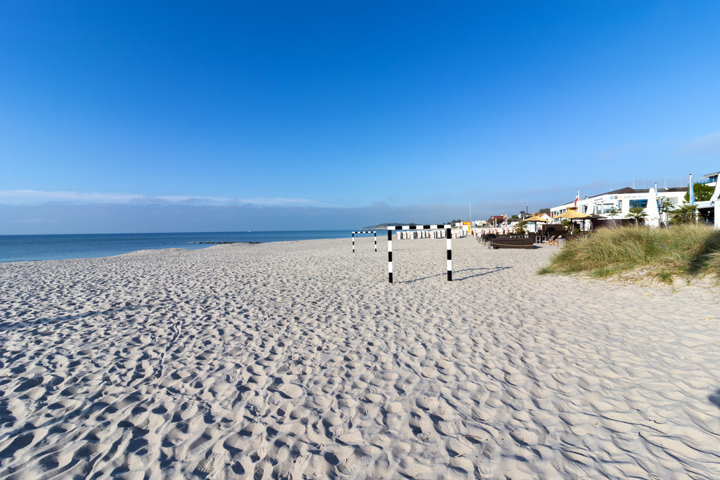 Wandbild: Zwischen Tor und Horizont – Küstenfreude pur | Dieses Wandbild zeigt eine sommerliche Strandszene in Grömitz. Der helle Sand erstreckt sich bis zum Horizont, durchzogen von Fußspuren, die von Bewegung und Lebendigkeit zeugen. Zwei schwarz-weiß gestreifte Tore stehen auf dem Strand – grafisch markant und zugleich Hinweis auf sportliche Aktivität, etwa Beachsoccer. Rechts im Bild befinden sich mehrere Gebäude mit Außenterrassen, darunter Strandcafés und Restaurants, die zum Verweilen einladen. Der Himmel ist wolkenlos und strahlt in klarem Blau, das Meer liegt ruhig im Hintergrund und schafft eine offene, entspannte Atmosphäre. Die Komposition lebt vom Wechselspiel aus Struktur und Weite, aus Bewegung und Ruhe – ein Moment der Küstenfreude. Dieses Motiv eignet sich ideal als Wandbild für maritime Wohnkonzepte – ob als Leinwandbild, Acrylglasbild, Alu-Dibond FineArt Print oder als Akustikbild. Ein stilvoller Akzent für Wohnzimmer, Büro oder Ferienwohnung. - Realisiert mit Pictrs.com