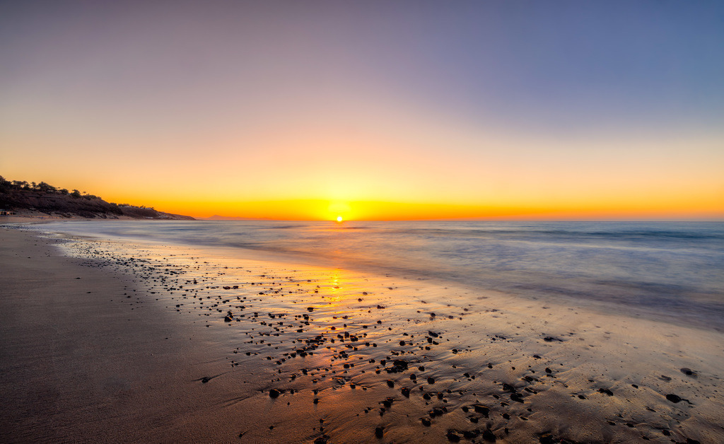 Sonnenaufgang über glitzernde Steine am Strand-Fuerteventura | Ein neuer Tag beginnt , am Strand auf Fuerteventura. Eine Aufnahme mit langer Belichtungszeit. Durch den tiefen Stand der Sonne, leuchten die feuchten Steine orange rot im funkelndem Licht der Sonne. - Realisiert mit Pictrs.com