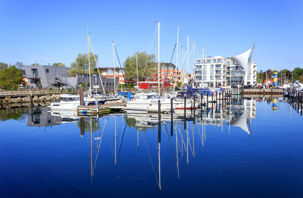 Wandbild: Spiegelung am Yachthafen in Eckernförde | Dieses Wandbild im Querformat zeigt den Yachthafen in Eckernförde. Auf dem Wasser im Vordergrund ist eine schöne Spiegelung der Segelboote sowie der Gebäude zu sehen. - Realisiert mit Pictrs.com