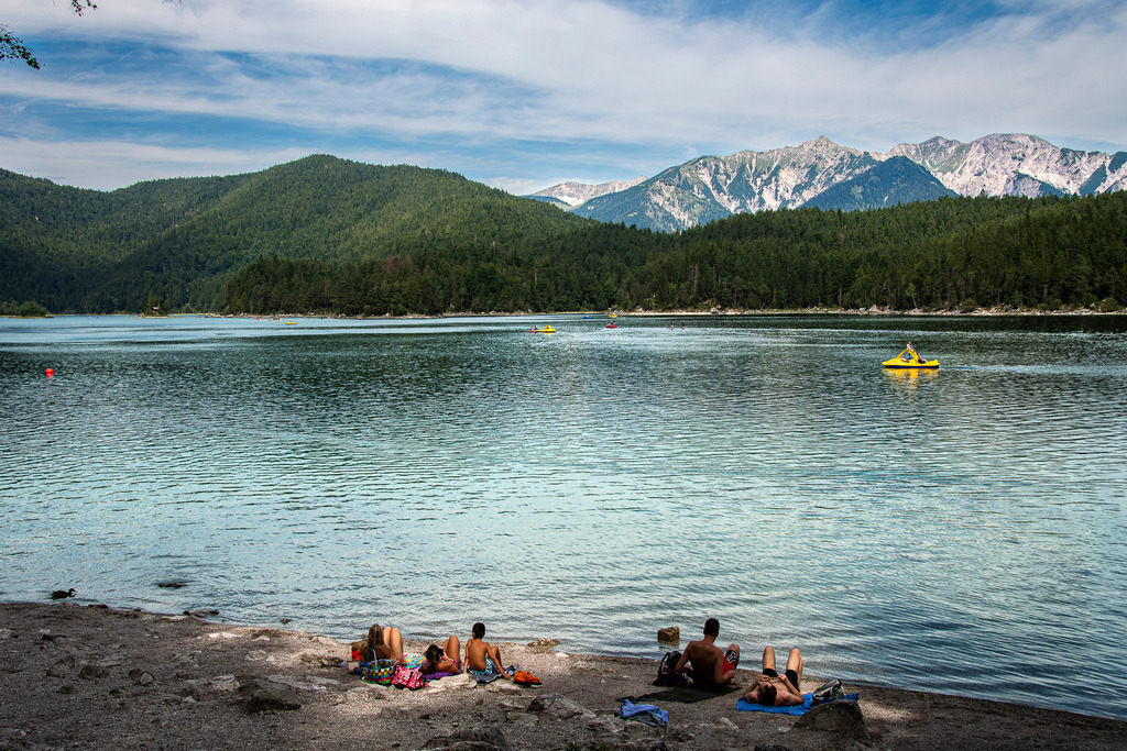 Sommerglück am türkisfarbenen Eibsee | Ein perfekter Sommertag am Eibsee nahe Garmisch-Partenkirchen: Badegäste entspannen am kiesigen Ufer und genießen die warme Sonne, während auf dem glasklaren, schimmernden Wasser ein gelbes Tretboot sanft seine Bahnen zieht. Im Hintergrund erheben sich bewaldete Hügel und die eindrucksvollen Gipfel des Wettersteingebirges unter einem leicht bewölkten Sommerhimmel. Die idyllische Szenerie strahlt Leichtigkeit, Urlaubsfreude und pure Erholung inmitten der majestätischen bayerischen Alpenwelt aus. - Realisiert mit Pictrs.com