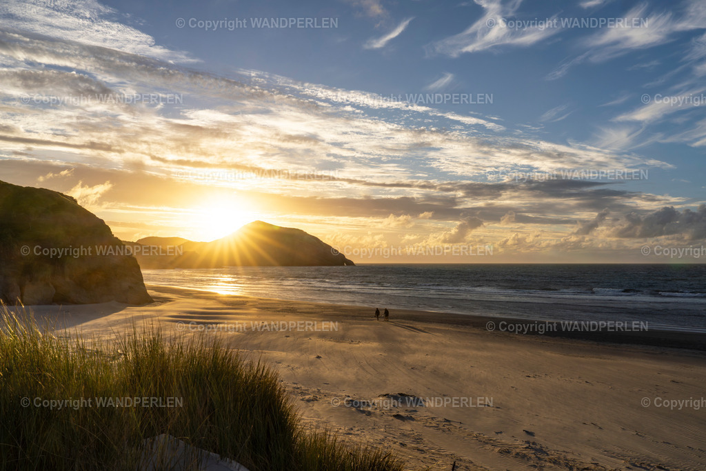 Goldener Sonnenuntergang am Wharariki Beach | Ein atemberaubender Sonnenuntergang beleuchtete den Wahariki Strand in Neuseeland und warf goldene Strahlen auf das Meer und den Sand. Zwei Silhouetten wandern entlang der Küste, umrahmt von felsigen Klippen und zarten Wolken. Die friedliche Szene betont die natürliche Schönheit dieser Bucht. - Realisiert mit Pictrs.com