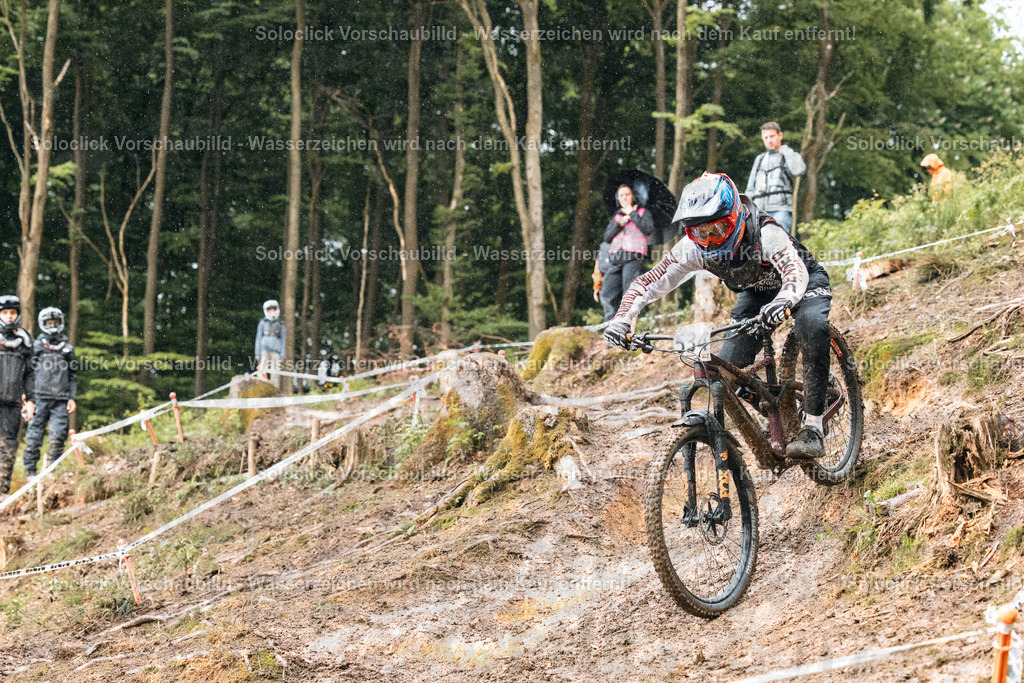 Downhill-Mountainbiker | Ein Mountainbiker, der einen steilen, verschneiten Hang hinunterfährt, umgeben von Bäumen und einem Zuschauerpublikum. Der Fahrer trägt einen Helm und Schutzkleidung und fährt ein Mountainbike mit Federgabel und Scheibenbremsen. Der Hang ist mit Schnee bedeckt und mit Markierungen versehen, was darauf hindeutet, dass es sich um einen Downhill-Kurs handelt. - Realisiert mit Pictrs.com
