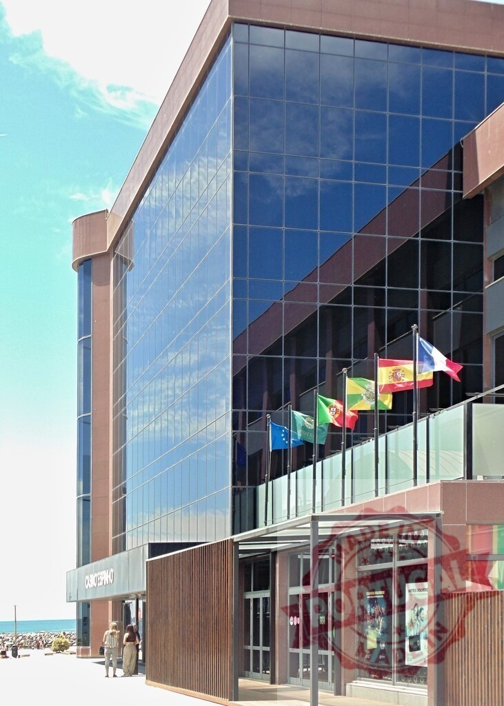 Casino Espinho modernist building with international flags. Aveiro - Portugal | Casino building featuring glass facade, flags, and reflections under a blue sky in Espinho - Realisiert mit Pictrs.com