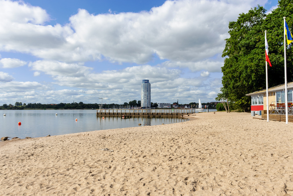 Wandbild: Schleistrand Luisenbad in Schleswig | Dieses Wandbild im Querformat zeigt den Schleistrand Luisenbad in Schleswig im Sommer. In der Ferne kann man den Wikingturm sehen. Am blauen Himmel befinden sich sommerliche Wolken.  - Realisiert mit Pictrs.com