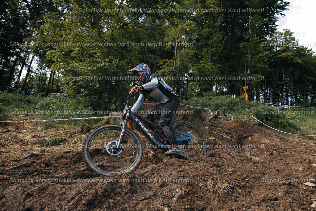Mountainbiker in Aktion | Ein Mountainbiker in voller Ausrüstung rast einen staubigen, schmalen Pfad in einem bewaldeten Gebiet hinunter. Der Fahrer trägt einen Helm, eine Brille und Schutzkleidung. Das Gelände ist steil und uneben, mit freiliegendem Erdreich und Wurzeln. Im Hintergrund sind Bäume und Büsche zu sehen. Die Szene vermittelt ein Gefühl von Geschwindigkeit und Abenteuer. - Realisiert mit Pictrs.com