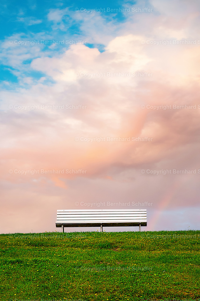 Sitzbank auf dem Deich | Eine weiße Sitzbank steht auf einem Deichhang an der Nordsee unter einem bewölkten Himmel mit Regenbogen. - Realisiert mit Pictrs.com