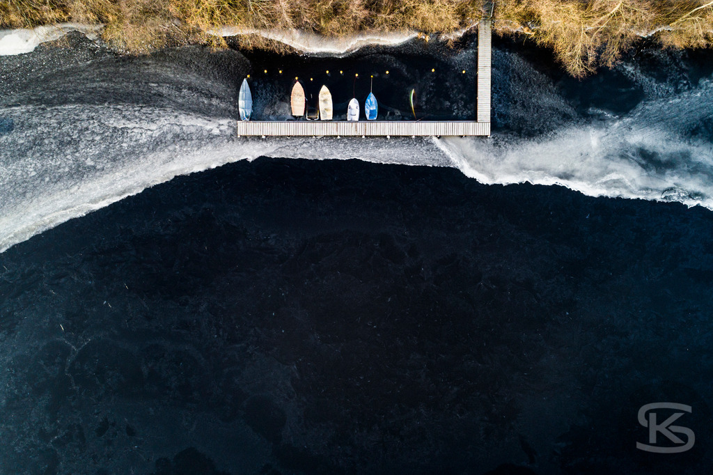 Eingefrorene Boote am Wintersteg – Drohnenaufnahme eines zugefrorenen Sees | Beeindruckende Luftaufnahme von eingefrorenen Booten am Steg eines zugefrorenen Sees. Die Drohnenfotografie zeigt winterliche Stille mit Baumschatten auf der Eisfläche aus der Vogelperspektive – atmosphärische Winterimpression. - Realisiert mit Pictrs.com