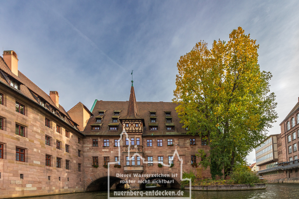 Monatsbild des Kalender 2026 für den Monat November | Heilig-Geist-Spital in Nürnberg, ein historisches Gebäude, das sich malerisch über die Pegnitz spannt. Im Vordergrund ist der Fluss zu sehen, der unter den Bögen des Gebäudes hindurchfließt. Rechts steht ein großer Baum, dessen Blätter in herbstlichen Farben leuchten. - Realisiert mit Pictrs.com