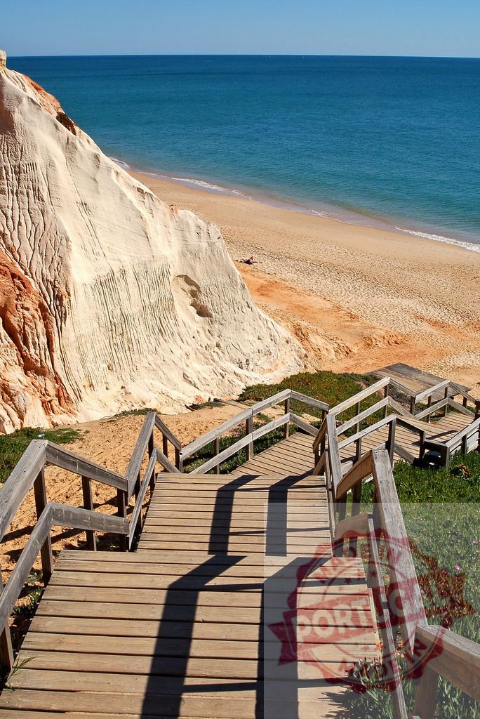 Wooden staircase leading down to sandy beach and ocean | Wooden staircase descending towards a golden sandy beach, eroded cliff, and blue Atlantic ocean - Realized with Pictrs.com