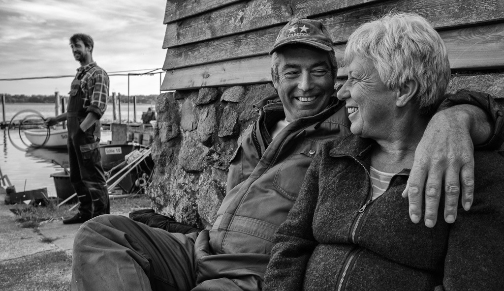 fischer-2020-094 | Den Feierabend genießt Jörn Ross, Berufsfischer in Schleswig an der Schlei, gerne mit seiner Frau Sabine auf einer Bank vor seinem Haus in Nienstadt, einer ruhigen Seitenstraße in der Fischersiedlung Holm. Im Hintergrund beobachtet Sohn Nils das Geschehen. Die Familie Ross lebt und fischt seit Jahrhunderten an der Schlei. - Realisiert mit Pictrs.com
