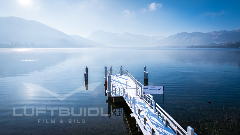 Gmund Kaltenbrunn; Schiffsanlgesteg; W36 | Der Schiffsanlegesteg in Gmund Kaltenbrunn liegt ruhig im klaren Wasser des Tegernsees und führt den Blick weit hinaus in die sanfte Weite der Landschaft. Zarte Nebelschleier und das ruhige Licht verleihen der Szene eine besondere Stille, während sich die umliegenden Berge nur schemenhaft am Horizont abzeichnen. Ein stiller Moment am See, der Gelassenheit und Weitblick auf besondere Weise verbindet. - Realisiert mit Pictrs.com