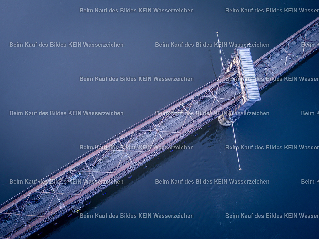 Hubbrücke Draufsicht-0057 | Das Luftbild zeigt die historische Hubbrücke in Magdeburg aus einer senkrechten Drohnenperspektive. Der Blick direkt von oben macht die markante Stahlfachwerkkonstruktion, die geometrische Linienführung und den Verlauf über die Elbe besonders eindrucksvoll sichtbar. Die außergewöhnliche Perspektive verdeutlicht die ingenieurtechnische Gestaltung des Bauwerks und seine Einbindung in die Flusslandschaft.Die Hubbrücke wurde 1848 als eingleisige Eisenbahnbrücke errichtet und zählt heute zu den ältesten erhaltenen Hubbrücken Deutschlands. In ihrer heutigen Form stammt die Stahlkonstruktion aus dem Jahr 1933. Das Bauwerk ist 220 Meter lang und besitzt ein 90 Meter langes bewegliches Mittelteil mit einem Gewicht von rund 450 Tonnen. Der Hubmechanismus konnte die Brücke innerhalb von etwa fünf Minuten um 2,87 Meter anheben, um auch bei Hochwasser die Schifffahrt auf der Elbe zu ermöglichen. Die senkrechte Drohnenaufnahme macht die technische Konstruktion der Hubbrücke in ihrer gesamten Dimension sichtbar. Das Zusammenspiel aus genieteter Stahlbauweise, Hubportalen und den klaren geometrischen Formen dokumentiert eindrucksvoll ein bedeutendes Denkmal deutscher Brücken- und Ingenieurbaukunst. - Realisiert mit Pictrs.com