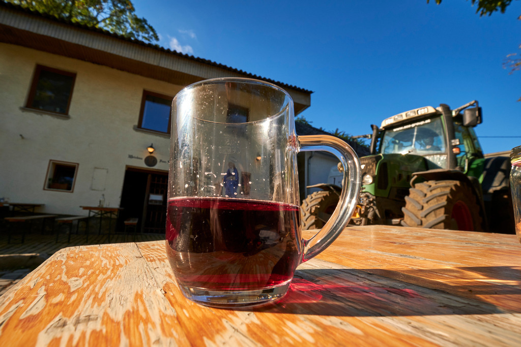 Ein Glas mit Traubensaft steht auf einem Holztisch | Pillichsdorf, Austria - October 16, 2021: Ein Glas mit Traubensaft steht auf einem Holztisch vor dem Kellerstöckl, im Hintergrund ein grüner Traktor. - Realisiert mit Pictrs.com