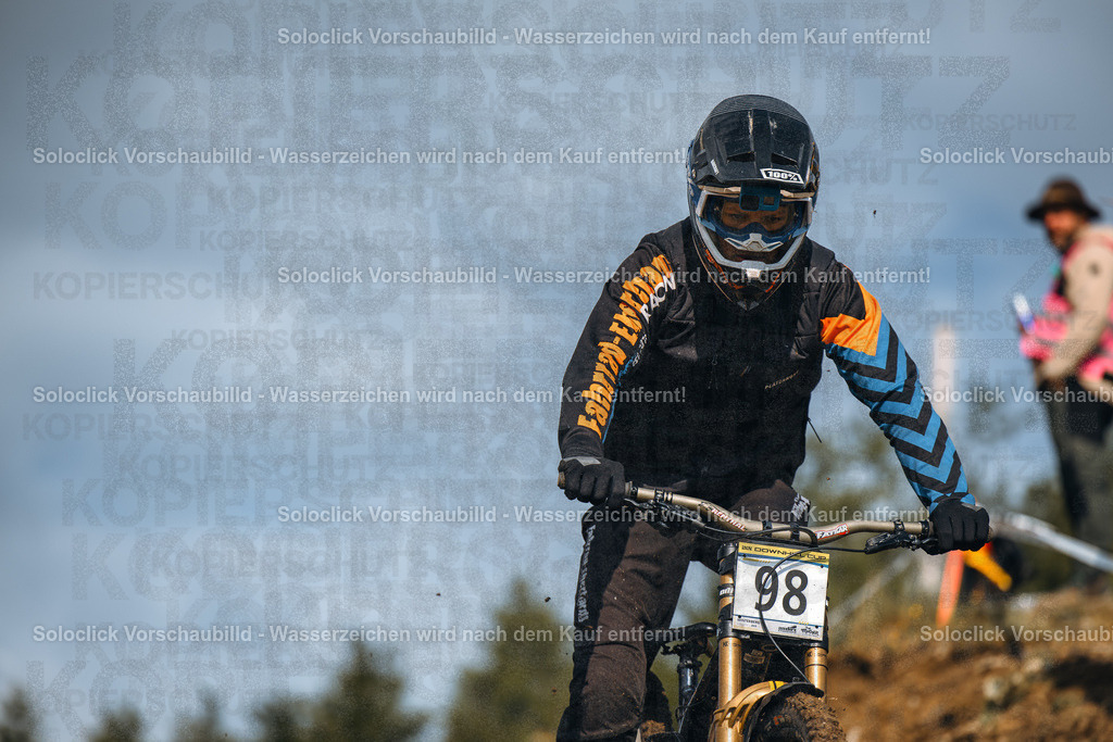 Mountainbiker in Aktion | Das Bild zeigt einen Mountainbiker in voller Montur, der einen staubigen, unebenen Pfad hinunterfährt. Er trägt einen schwarzen Helm mit dem Logo '100%' und eine dunkle Schutzausrüstung mit blauen und orangefarbenen Akzenten. Er trägt eine Nummer 98 auf seinem Rücken. Der Hintergrund besteht aus Bäumen und einem bewölkten Himmel. - Realisiert mit Pictrs.com
