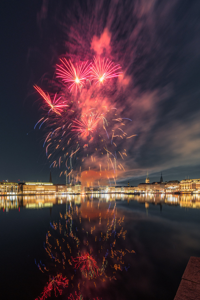 Rotes Feuerwerk über Hamburg bei Nacht | Rote und orange Feuerwerkslichter breiten sich über der Hamburger Alster aus und spiegeln sich im Wasser. Im Hintergrund leuchtet die nächtliche Innenstadt. - Realisiert mit Pictrs.com