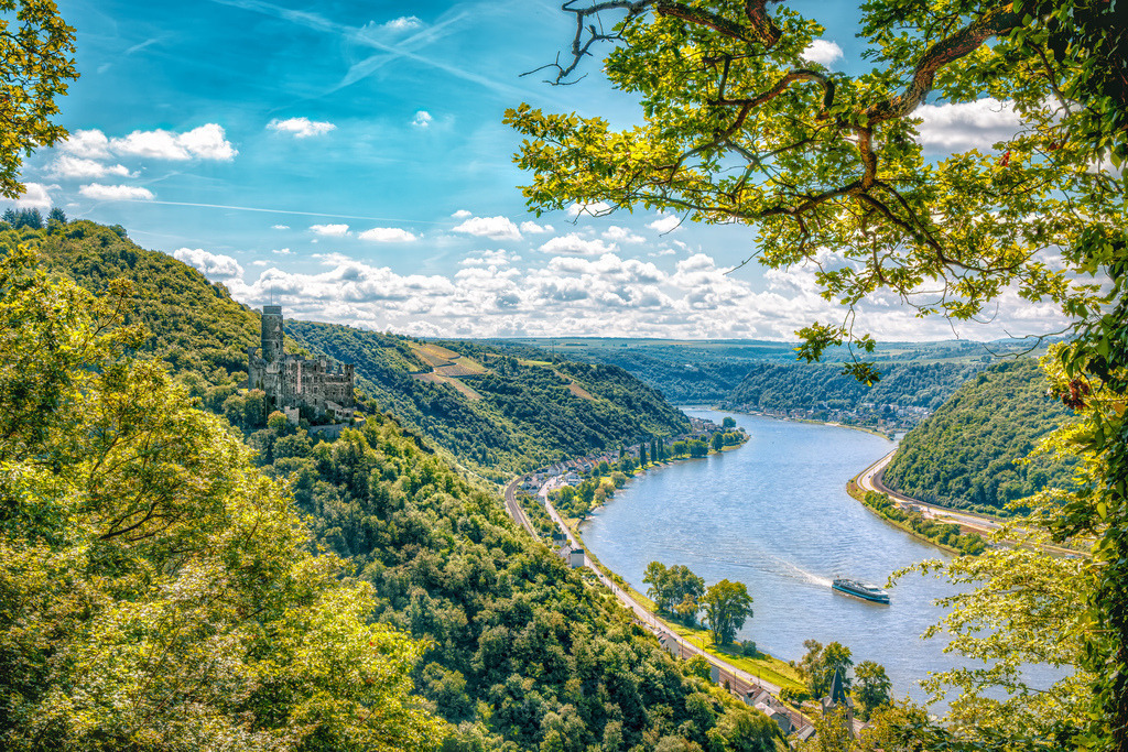 Panoramablick auf die malerische Rheinschleife mit Burganlage | Das Bild zeigt den Blick auf das UNESCO-Welterbe Oberes Mittelrheintal mit einer Rheinschleife und einer historischen Burganlage am bewaldeten Hang.Das Obere Mittelrheintal in Rheinland-Pfalz/Hessen, bekannt für seine Flusslandschaften, Steilhänge und zahlreichen Burgen.Der gewendelte Verlauf des Rheins, dichte Laubwälder an den Hängen sowie mittelalterliche Wehr- und Burgenarchitektur (wie typisch für den Rheinabschnitt um die Loreley oder Oberwesel) - Realisiert mit Pictrs.com