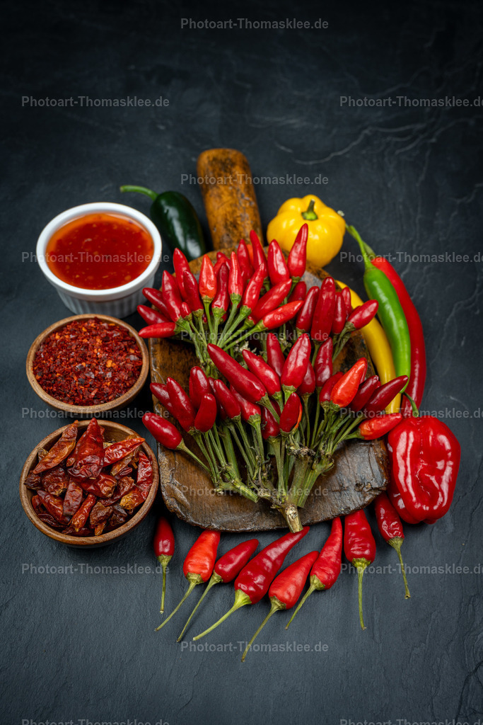 Scharfe Chilis mit Gewürzen auf dunklem Schiefer | Auf dunklem Schiefer arrangiert sich eine beeindruckende Sammlung scharfer Gewürze und Chilis. Leuchtend rote Chilischoten bilden das Zentrum der Komposition, umgeben von verschiedenen Gewürzschalen mit Chilipulver und getrockneten Chilis. Eine weiße Schale enthält rote Chilisauce, während die alte Gewürzschaufel und bunte Paprika die Szene ergänzen. Der dunkle Hintergrund lässt die intensiven Rottöne der Chilis besonders leuchten. Diese professionelle Anordnung zeigt die ganze Vielfalt scharfer Küchengewürze in ihrer appetitlichen Pracht. - Realisiert mit Pictrs.com