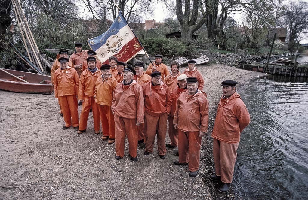 zeitenwende-farbe-02-01 | 1989 versammelten sich alle Mitglieder der Holmer Fischerzunft in Schleswig für dieses Gruppenbild. Ganz rechts: Harald Ross, der 1. Ältermann. Mit der Hand an der historischen Zunftfahne: Jörn Ross. Fünfter von rechts: Matthias Nanz. - Realisiert mit Pictrs.com