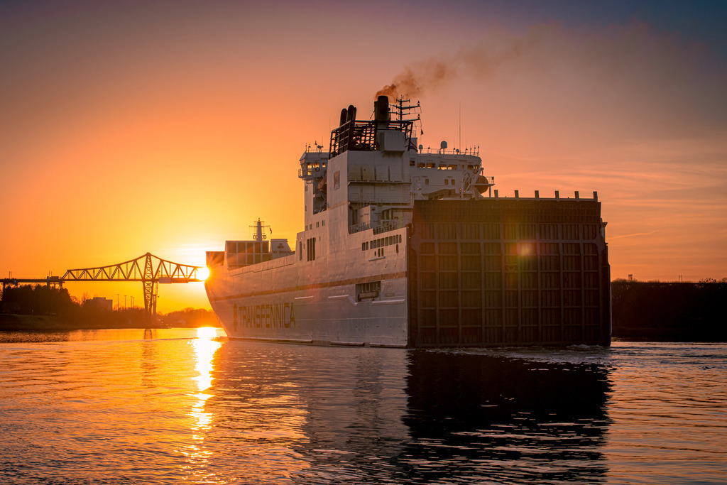 Rendsburger Hochbrücke im Sonnenuntergang | Eine LKW Fährschiff bewegt sich im Nord Ostsee Kanal langsam auf die majestätische Rendsburger Hochbrücke zu, während die Sonne sich langsam hinter dem Horizont senkt. Das glitzernde Wasser des Kanals spiegelt das orange-rote Licht des Sonnenuntergangs wider und verleiht der Szenerie eine fast magische Atmosphäre. Diese friedliche Stimmung der Szenerie lädt ein zum Innehalten.