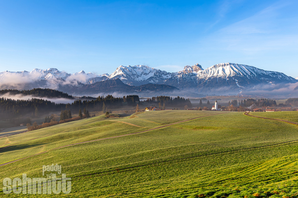 St. Moritz Dorfkirche bei Zell vor verschneiter Bergkulisse im Allgäu | Die Dorfkirche St. Moritz bei Zell im Allgäu vor der beeindruckenden Bergkulisse im frischen Frühlingslicht. Eine stimmungsvolle Landschaftsaufnahme mit typisch Allgäuer Charakter. - Realisiert mit Pictrs.com