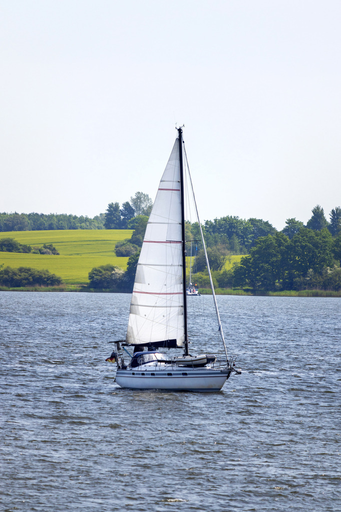 Wandbild: Segelboot auf der Schlei in Lindaunis | Dieses Wandbild im Hochformat zeigt ein Segelboot auf der Schlei im Hochformat in Lindaunis. Im Hintergrund ist frisches Frühlingsgrün zu sehen.  - Realisiert mit Pictrs.com