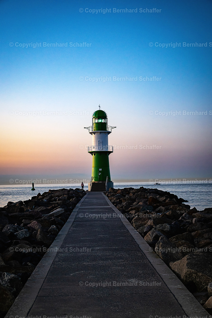 Leuchtturm im Abendlicht | Grüner Leuchtturm und Steinküste mit Fußweg an der Hafeneinfahrt von der Ostsee nach Warnemünde / Rostock in Deutschland. Abendstimmung bei Sonnenuntergang zur blauen Stunde und eine Person blickt auf das offene Meer. - Realisiert mit Pictrs.com
