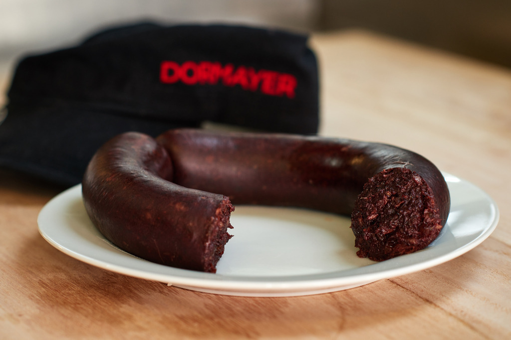 Fleischerei Dormayer | Langenzersdorf, Austria - February 12, 2019: Fleischerei Dormayer - Blutwurst Herstellung; angeschnittene Blutwurst auf einem Porzellanteller, im Hintergrund eine schwarze Kappe mit Logo. - Realisiert mit Pictrs.com