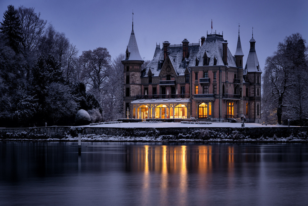 Winterliches Schloss Schadau am Thunersee | Das historische Schloss Schadau präsentiert sich in einer winterlichen Morgendämmerung. Die warmen Lichter aus den Fenstern des Schlosses spiegeln sich deutlich auf der ruhigen Wasseroberfläche im Vordergrund. Eine Schneedecke bedeckt das Dach und die umliegenden Bäume, was eine stimmungsvolle Szenerie schafft. - Realisiert mit Pictrs.com