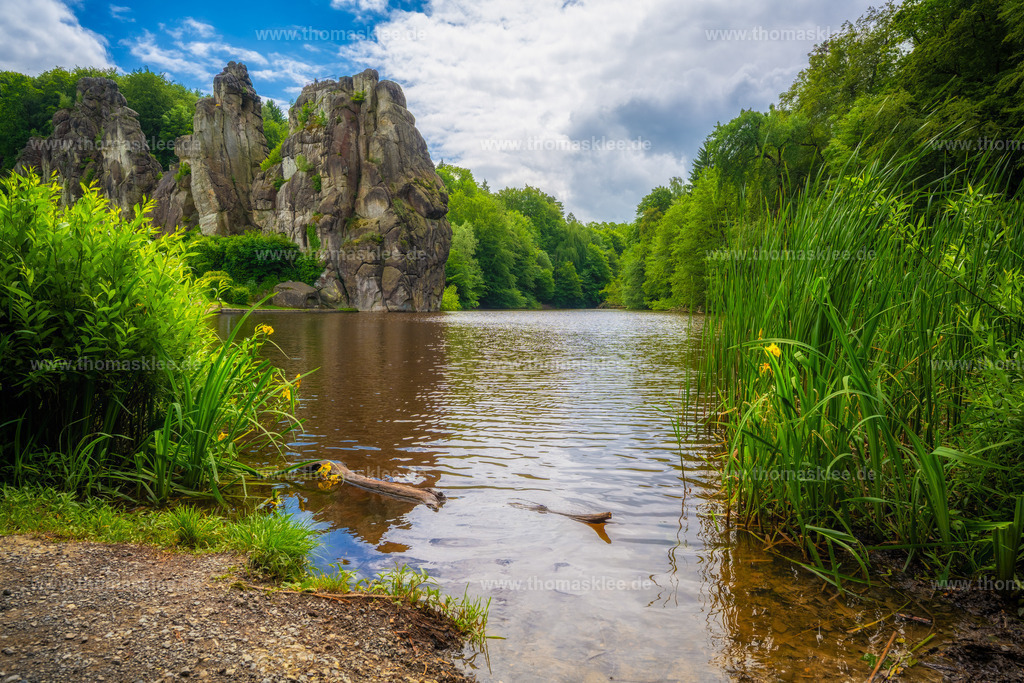 Externsteine – Natur pur am stillen Waldsee | Dieses Bild nimmt einen auf eine andere Art mit als die klassischen Panoramaaufnahmen der Externsteine — es ist näher dran, lebendiger, fast schon zum Anfassen. Der Blick führt vom Ufer des kleinen Sees im Vordergrund direkt auf die mächtigen Sandsteinfelsen im Hintergrund, die sich hoch über das Grün der Bäume erheben. Rechts säumen hohe Schilfrohre das Ufer, links wachsen gelb blühende Wildpflanzen ins Bild. Ein alter, verwitterter Ast liegt halb im Wasser — ein kleines, unscheinbares Detail, das dem Ganzen eine echte, ungekünstelte Natürlichkeit gibt.Die Farben sind frisch und lebendig: sattes Grün in allen Schattierungen, das warme Braun des seichten Wassers, der helle Kies am Uferrand und ein freundlicher Himmel mit lockeren Wolken darüber. Es wirkt wie ein ruhiger Sommertag — man meint fast, das leichte Plätschern des Wassers zu hören.Die Externsteine selbst haben eine beeindruckende Geschichte. Diese Sandsteinformation im Teutoburger Wald in Nordrhein-Westfalen gilt als eines der bedeutendsten Naturdenkmäler Deutschlands. Bereits in vorchristlicher Zeit war dieser Ort ein Heiligtum, später wurden Kapellen und Reliefs direkt in den Fels gehauen. Diese tiefe historische Verwurzelung schwingt auch in diesem Bild mit — auch wenn die Aufnahme auf den ersten Blick einfach nur schön und leicht wirkt.Fotografisch lebt das Bild von seiner bodennahen Perspektive und dem starken Vordergrundinteresse. Statt den Fels von Weitem zu zeigen, führt der Fotograf den Betrachter mitten ins Geschehen. Das Ergebnis ist eine Aufnahme, die sich frisch und authentisch anfühlt — kein aufgesetztes Postkartenmotiv, sondern ein echter Moment in der Natur. - Realisiert mit Pictrs.com