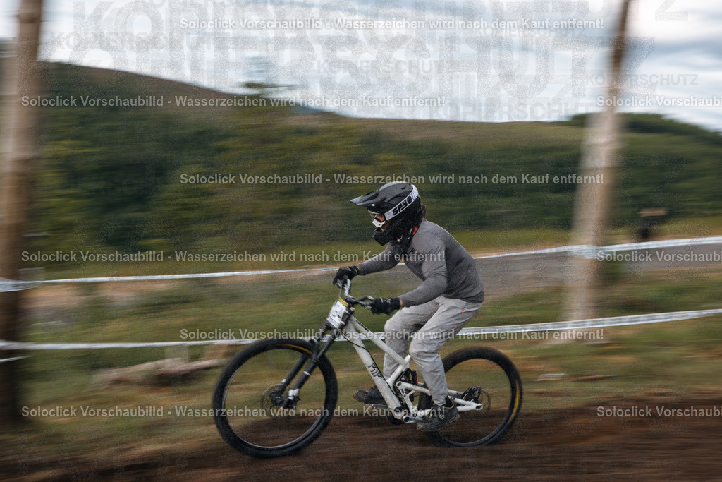 Geschwindigkeit und Präzision: Downhill-Action im Bikepark Winterberg | Ein Mountainbiker, vollständig in schwarzer Schutzausrüstung gekleidet, rast mit hoher Geschwindigkeit einen staubigen, schmalen Pfad hinunter. Der Fokus liegt auf dem Fahrer und seinem Mountainbike, während der Hintergrund verschwommen ist, was die Geschwindigkeit und Bewegung betont. Der Fahrer trägt einen schwarzen Helm mit der Aufschrift 'SPV', Handschuhe und eine dunkle Jacke und Hose. Das Mountainbike ist ein Downhill-Bike mit dicken Reifen und einer robusten Federgabel. Der Pfad schlängelt sich durch eine offene Landschaft mit vereinzelten Bäumen und einer staubigen Oberfläche. - Realisiert mit Pictrs.com