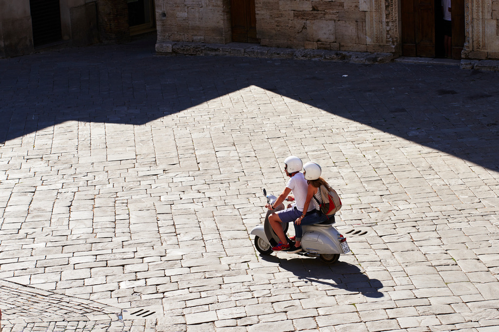 Junges Paar auf einer grauen Vespa | Bevagna, Italien - August 28, 2016: Junges Paar auf einer grauen Vespa fährt über den Dorfplatz. - Realisiert mit Pictrs.com