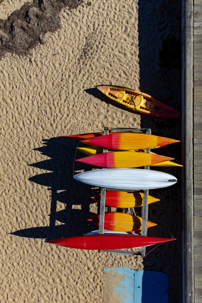 Shades of Summer | From above, the rhythm of color unfolds — kayaks in bright reds, oranges, and yellow tones rest quietly on the sand. A moment of geometric harmony where summer light meets seaside calm. - Realisiert mit Pictrs.com