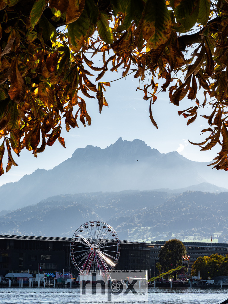 Pilatus. Herbst. Riesenrad. | <p><strong>FARBE. GEBIRGE. HORIZONT.</strong></p><p>Die Herbstpracht des Kastanienlaubs bildet einen dramatischen Rahmen für den majestätischen Pilatus über dem Vierwaldstättersee. Im Vordergrund setzt das leuchtende Riesenrad einen modernen Akzent zur zeitlosen Schönheit der Alpen. Dieses Bild fängt den einzigartigen Kontrast zwischen städtischem Leben und alpiner Erhabenheit ein.</p><p>Wähle unter "Produktauswahl" dein Wunschformat: Vom klassischen Wandbild über Puzzle & Tassen bis zum digitalen Download (z.B. als Handy-Hintergrund).</p> - Realisiert mit Pictrs.com