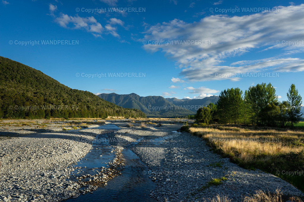 Maruia River im goldenen Nachmittagslicht | Ein ruhiges, teilweise trockenes, naturbelassenes Flussbett schlängelt sich durch ein Tal, flankiert von dichtem Wald und hohen Bergketten unter einem leuchtend blauen Himmel mit zerstreuten weißen Wolken. Die Szene, am späten Nachmittag aufgenommen, betont die rauhe Schönheit der Südalpen Neuseelands, wobei das Sonnenlicht lange Schatten auf den Kies und die spärliche Vegetation wirft. - Realisiert mit Pictrs.com