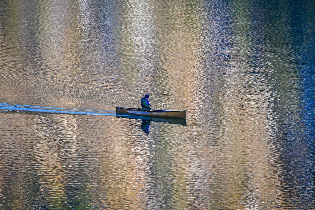 Im Einklang mit der Stille | Ein winziges Boot auf einer schier endlosen, golden schimmernden Leinwand: Sanfte Wellen brechen das warme Licht und verwandeln das Wasser in flüssiges Gold. Mitten in dieser gewaltigen Kulisse gleitet ein Kanufahrer lautlos dahin. Eine feine Spur im Wasser erzählt von seiner Reise, während sich die Welt um ihn herum in tiefer Ruhe verliert. Es ist dieser eine, kostbare Moment absoluter Einsamkeit – ein stiller Dialog zwischen Mensch und Natur, der den Lärm des Alltags augenblicklich verstummen lässt. - Realisiert mit Pictrs.com