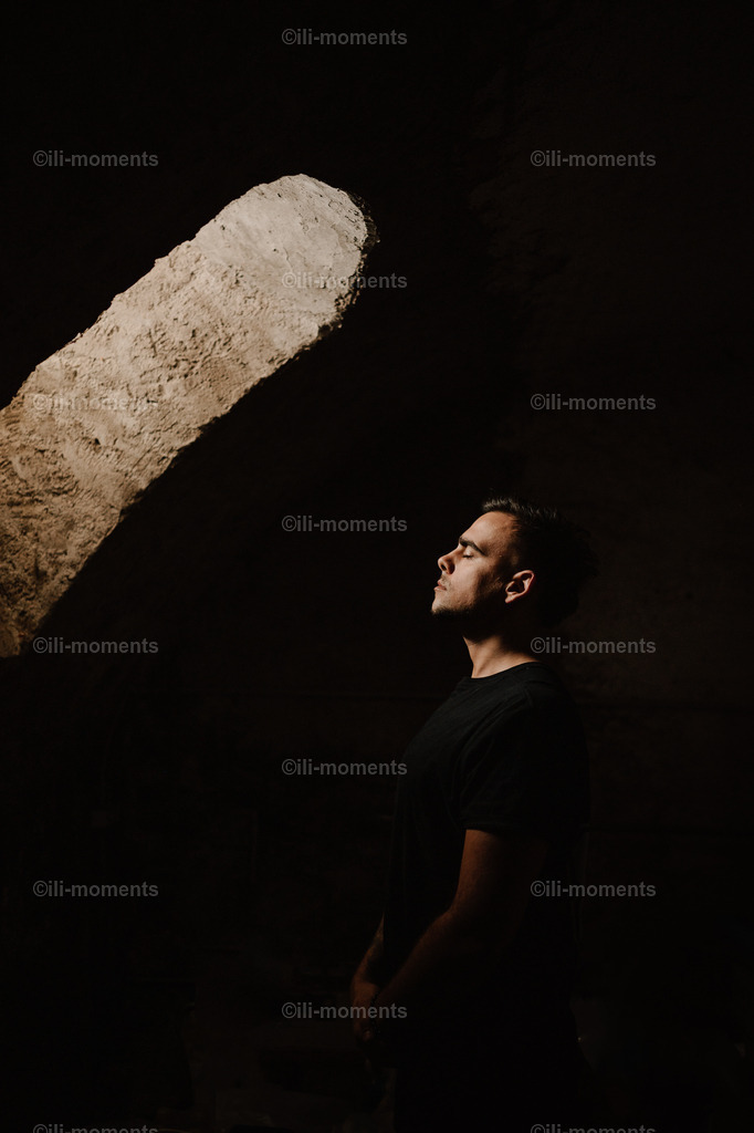 das einzige Licht | In einem dunklen Raum fällt Sonnenlicht durch ein kleines burgenähnliches Fenster auf das Gesicht eines Mannes mit geschlossenen Augen. Das Bild strahlt Ruhe, Genuss und Zufriedenheit aus – ideal als stimmungsvolle Fotokunst. - Realisiert mit Pictrs.com
