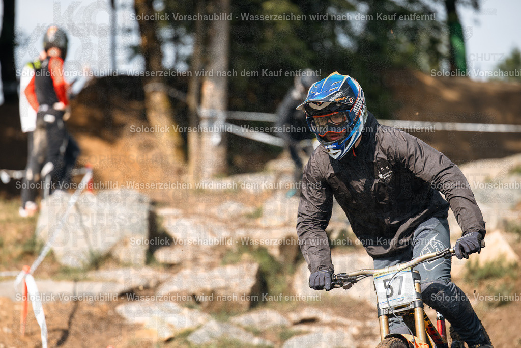 Downhill-Mountainbiker in Aktion | Ein Mountainbiker in voller Ausrüstung rast einen staubigen, steilen Pfad hinunter. Er trägt einen blauen Helm mit einer Visier und eine dunkle Jacke und Hose. Er hält ein Mountainbike mit roten und schwarzen Akzenten. Im Hintergrund sind weitere Mountainbiker und eine steile, erdige Klippe zu sehen. - Realisiert mit Pictrs.com