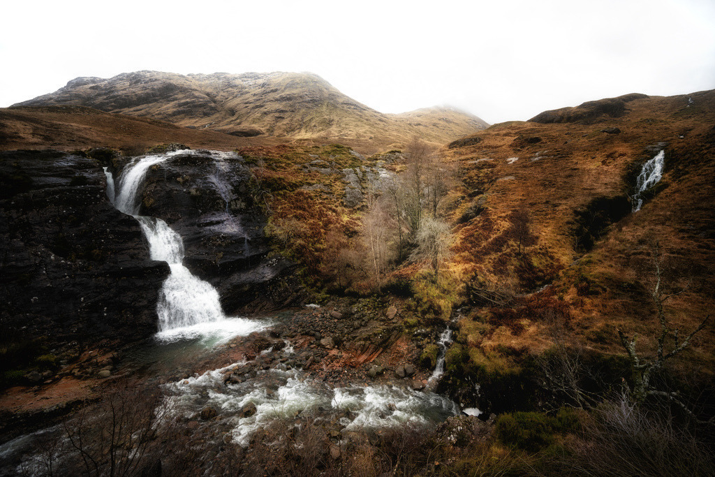 Glencoe Wasserfall, Schottland | Ein majestätischer Wasserfall stürzt sich über dunkle Felsen in ein rauschendes Flussbett, umgeben von den weitläufigen, braun-orangefarbenen Hängen der schottischen Highlands. Die winterliche Landschaft von Glencoe wird von einem bedeckten Himmel dominiert, der die natürliche, raue Schönheit der Region hervorhebt. Die Komposition betont die vertikalen Linien des Wasserfalls und die horizontalen Strukturen der Berge. - Realisiert mit Pictrs.com