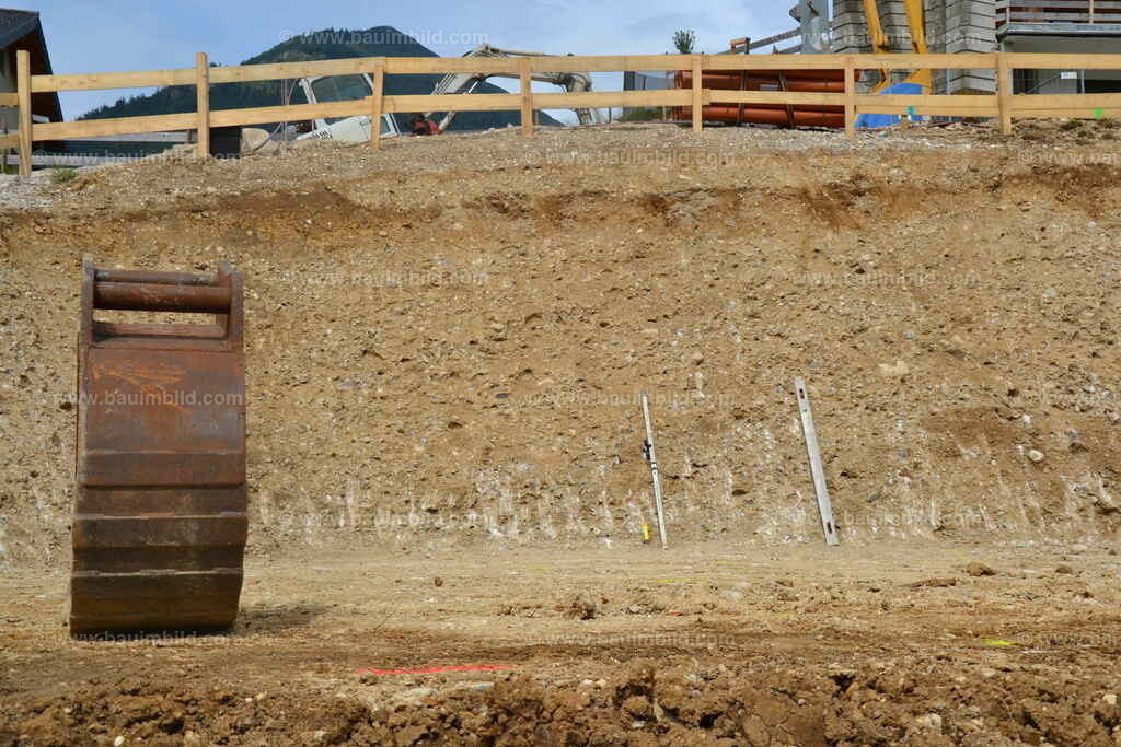 Baugrubenwand | Baugrube in Hanglage mit steil geböschter Baugrubenwand in Schotterboden, Bodenklasse 4, oberhalb Absturzsicherung mit Holzzaun, Baggerlöffel auf Baugrubensohle im Vordergrund