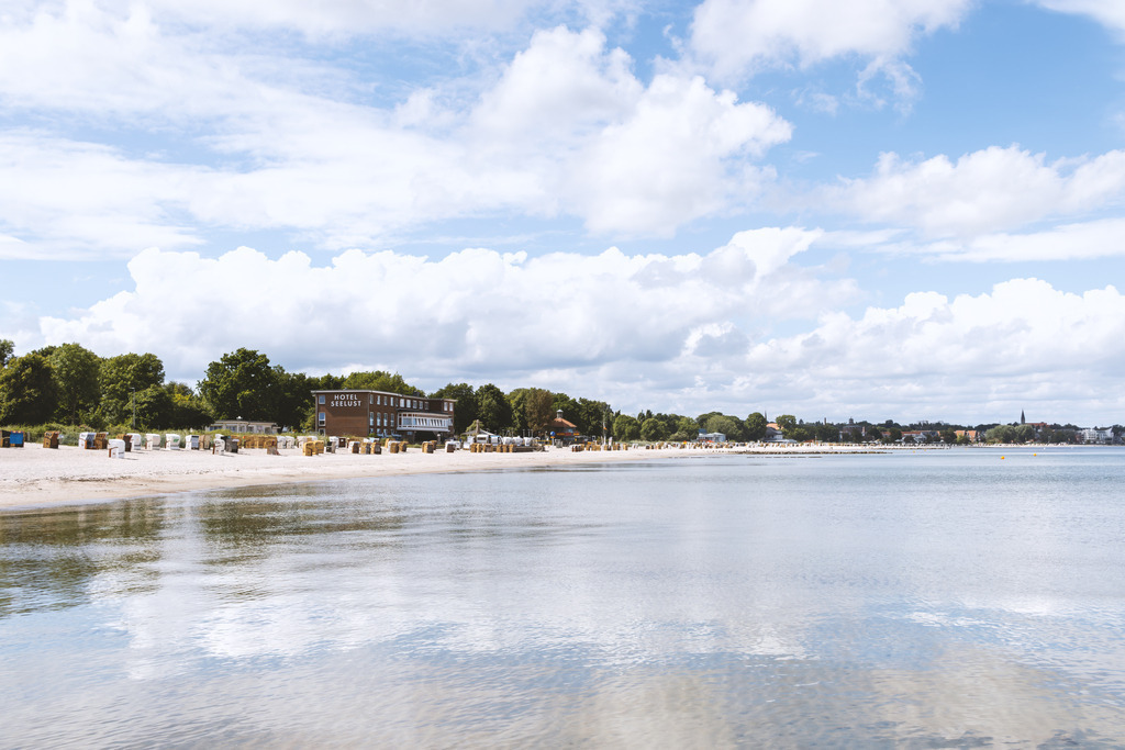 Wandbild: Küstenruhe von der Meerseite – Eckernförde im sanften Licht | Der Strand von Eckernförde zeigt sich aus der Perspektive der Meerseite mit seinen geordneten Strandkörben, dem hellen Sand und dem Hotel Seelust im Hintergrund. Die ruhige Wasseroberfläche im Vordergrund und der weite Himmel verleihen dem Motiv eine entspannte, sommerliche Atmosphäre. Die Kombination aus Küstenlandschaft, maritimer Architektur und der sanften Weite des Horizonts schafft ein authentisches Bild der Ostseeküste, das die besondere Stimmung von Eckernförde eindrucksvoll einfängt.