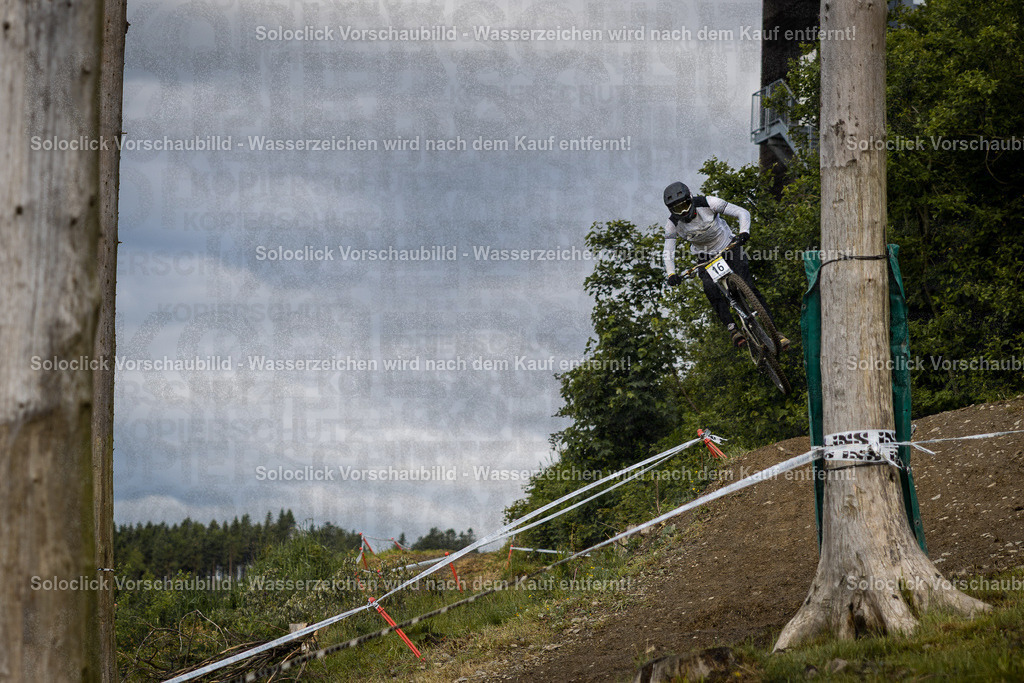 Action im Bikepark Winterberg | Das Foto zeigt einen Mountainbiker in voller Ausrüstung, der während des iXS Downhill Cups 2025 im Bikepark Winterberg, speziell auf der Strecke 'The Mother', einen Sprung vollführt. Er trägt einen weißen Helm mit der Nummer 16, eine dunkle Brille und eine dunkle Jacke. Der Hintergrund besteht aus bewaldete Hügel und ein trüber Himmel. Die Strecke ist mit roten und weißen Markierungen versehen. - Realisiert mit Pictrs.com