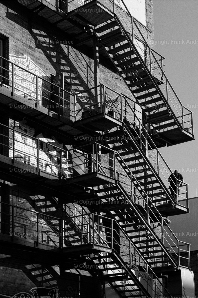 Abendsonne Berlin Prenzlauer Berg | Ein Mann steht auf einer Außentreppe bei tiefstehender Sonne. Straßenfotografie aus Berlin in schwarz weiß. - Realisiert mit Pictrs.com