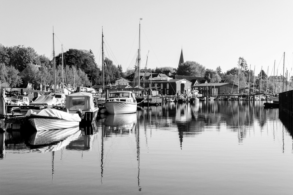 Wandbild: Hafen in Eckernförde in Schwarz-Weiß | Dieses Wandbild im Querformat zeigt den  Hafen in Eckernförde in Schwarz-Weiß. Im Vordergrund ergibt sich eine schöne Spiegelung im Wasser. Der Himmel ist wolkenlos.  - Realisiert mit Pictrs.com