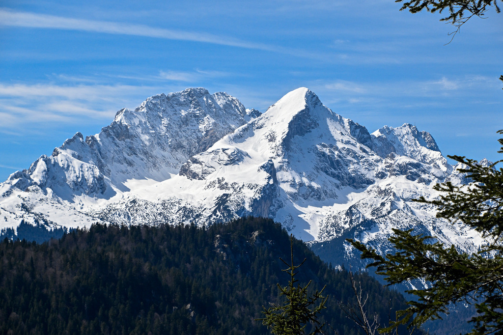 Der Ruf der Freiheit | Ein Anblick, der das Herz höherschlagen lässt: Stolz und majestätisch erhebt sich die verschneite Alpspitze neben dem mächtigen Zugspitzmassiv in den strahlend blauen Himmel, sanft durchzogen von weißen Schleierwolken. Unterhalb der schroffen, eisigen Riesen erstreckt sich das tiefe Grün der dichten Nadelwälder der Region Garmisch-Partenkirchen. Dieses Bild fängt die pure, ungezähmte Kraft der Alpen ein – ein stilles Monument der Natur im Zugspitzgebiet, das Sehnsucht nach Abenteuer und innerem Frieden weckt. - Realisiert mit Pictrs.com