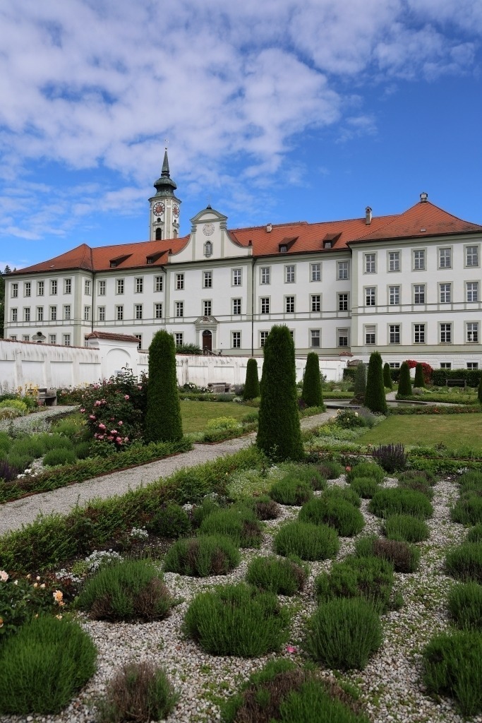 Prälatengarten am Kloster Schäftlarn | Das Foto zeigt den gepflegten Prälatengarten des Kloster Schäftlarn. Die Anlage ist durch klare Formen, symmetrische Wege und sorgfältig angelegte Beete geprägt. Üppige Blumenrabatten, niedrige Hecken und kunstvoll geschnittene Sträucher verleihen dem Garten eine elegante, barock inspirierte Ausstrahlung.Zwischen den Beeten führen gerade Wege durch die Anlage und lenken den Blick auf die harmonische Gesamtgestaltung. Im Hintergrund erhebt sich die historische Klosteranlage mit ihrer beeindruckenden Architektur, die den Garten als würdigen Rahmen ergänzt. Das frische Grün der Pflanzen bildet einen reizvollen Kontrast zu den hellen Fassaden und den warmen Farbtönen der Gebäude.Das Sonnenlicht hebt die feinen Strukturen der Bepflanzung hervor und lässt die Farben der Blüten lebendig erscheinen. Die Atmosphäre des Fotos wirkt ruhig, geordnet und besinnlich. Der Prälatengarten vermittelt die Verbindung von klösterlicher Tradition, gärtnerischer Kunst und der Schönheit der Natur und lädt zum Verweilen und Genießen ein. - Realisiert mit Pictrs.com