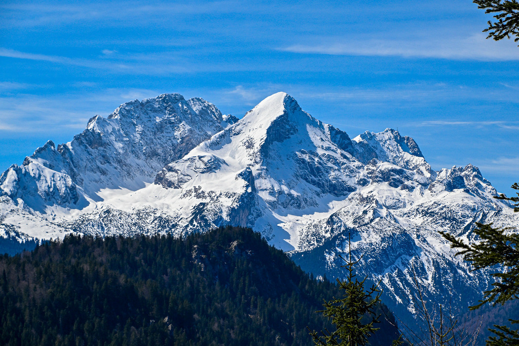 Im Bann der weißen Riesen | Ein überwältigender Blick auf das Herz des Alpenraums: Stolz und formvollendet erhebt sich die pyramidenförmige Alpspitze im wunderschönen Werdenfelser Land. Flankiert vom mächtigen Zugspitzmassiv und unweit von Deutschlands höchstem Berg, der Zugspitze, strahlt dieses alpine Panorama eine zeitlose Magie aus. Unter dem intensiv blauen Himmel ruhen die dunklen Nadelwälder der Region Garmisch-Partenkirchen wie ein schützendes Band. Ein Bild voller Urkraft und unendlicher Weite, das die Seele tief im Zugspitzgebiet zur Ruhe kommen lässt. - Realisiert mit Pictrs.com