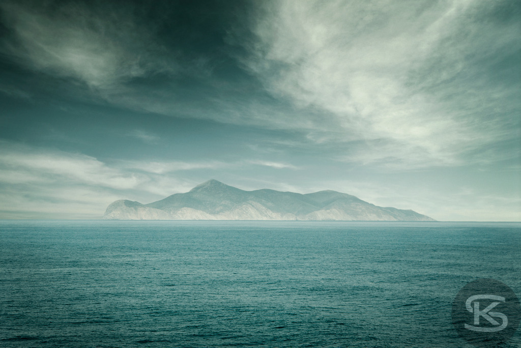 Neblige Insel am Horizont – Ruhige Meereslandschaft mit Bergen | Stimmungsvolle Meereslandschaft mit einer nebligen Insel und Bergsilhouette am Horizont. Ruhige See, dramatischer Himmel und eine minimalistische, zeitlose Atmosphäre. - Realisiert mit Pictrs.com