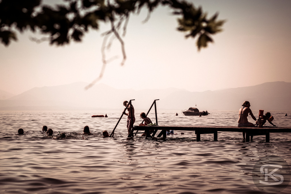 Stimmungsvolle Badeszene am Gardasee bei Sonnenuntergang | Ein idyllisches Bild einer Badeszene am Gardasee. Das warme, weiche Licht des Sonnenuntergangs beleuchtet das Wasser und die umliegenden Berge, während Menschen in Silhouette am Steg sitzen und schwimmen. Die Aufnahme fängt die friedliche und nostalgische Urlaubsatmosphäre perfekt ein. - Realisiert mit Pictrs.com
